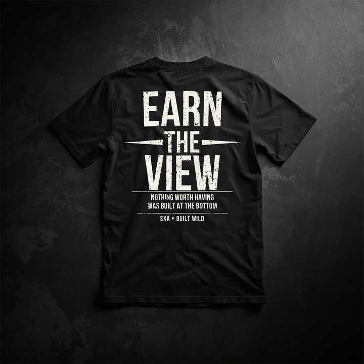 An image of the back of a black t-shirt featuring a distressed white graphic design. The central text reads "EARN THE VIEW" in a bold, weathered sans-serif font. The word "THE" is flanked by two horizontal sharp, needle-like lines. Below this, a smaller slogan reads, "NOTHING WORTH HAVING WAS BUILT AT THE BOTTOM," framed by two thin horizontal lines. At the very bottom, the branding "SXA • BUILT WILD" is displayed. The shirt is laid flat against a dark, textured slate-like background.