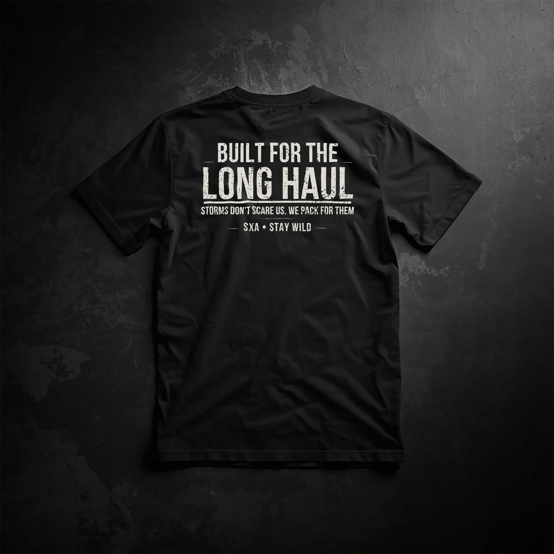 An image of the back of a black t-shirt featuring a white distressed graphic design. The primary text reads "BUILT FOR THE LONG HAUL" in a bold, weathered block font. Directly below, a smaller motivational slogan says, "STORMS DON’T SCARE US. WE PACK FOR THEM," followed by the brand signature "SXA • STAY WILD" centered between two thin horizontal lines. The shirt is displayed on a dark, textured charcoal background, emphasizing a rugged, outdoor adventure and overlanding aesthetic.